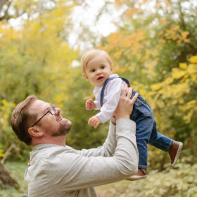 Father Lifting Baby Boy in Air Against Tree Foliage
