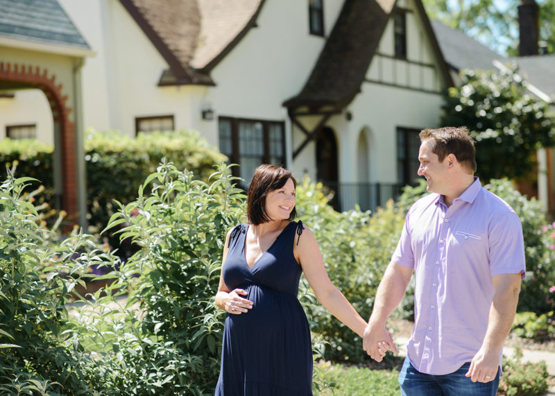 Pregnant couple holding hands and smiling in front of their home