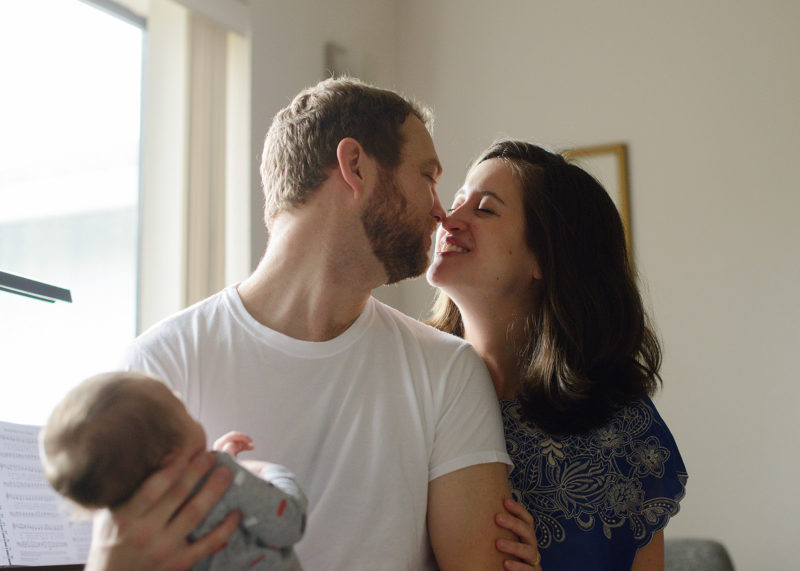 Mom and dad share a kiss in home while holding newborn baby