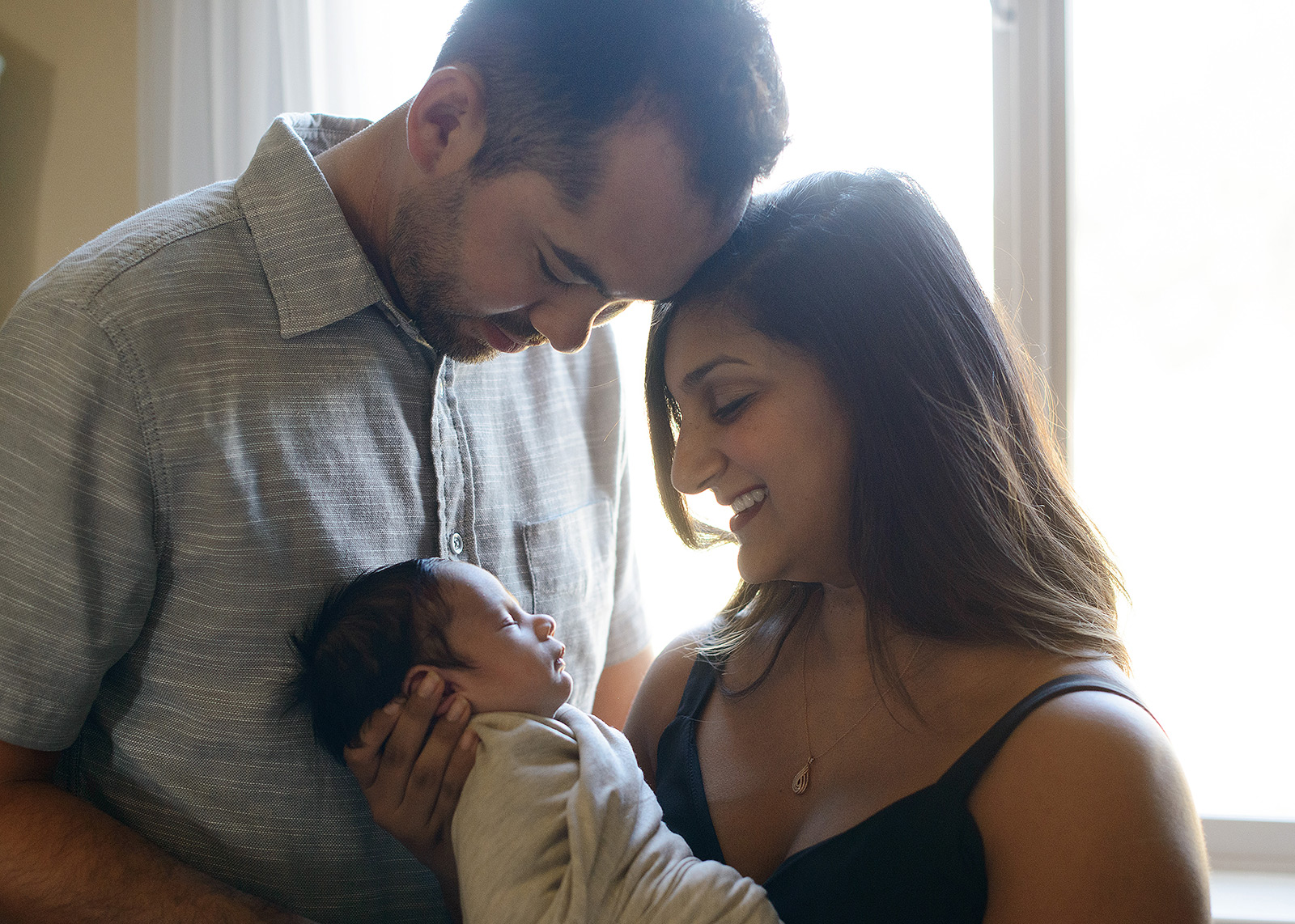 Mom and dad look lovingly at newborn sleepy baby against light in home