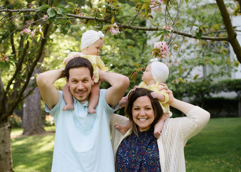 Family photo with twins on mom and dad's shoulders underneath cherry blossom tree