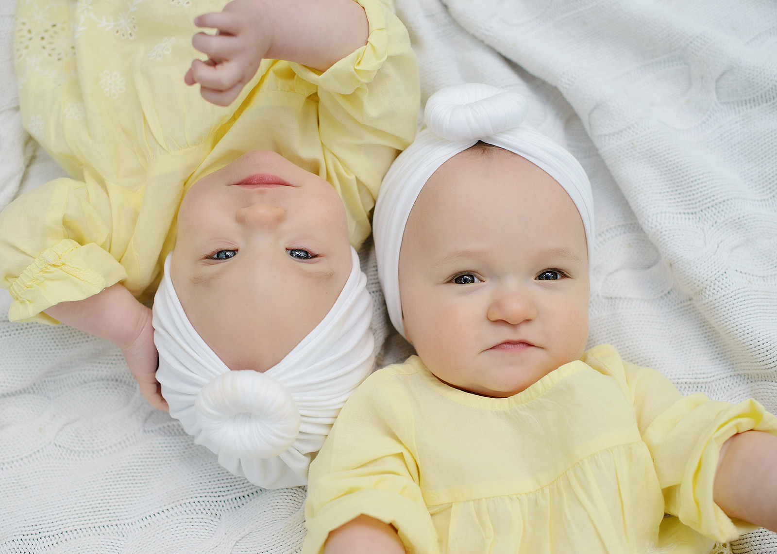 Twin baby girls wearing yellow lying down on white blanket and looking directly at camera