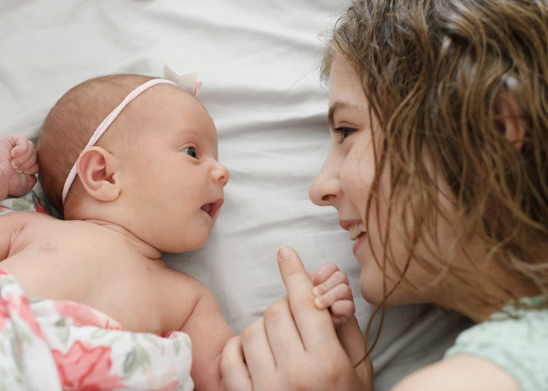 Big sister holds newborn baby sister's hand and stares lovingly into her eyes