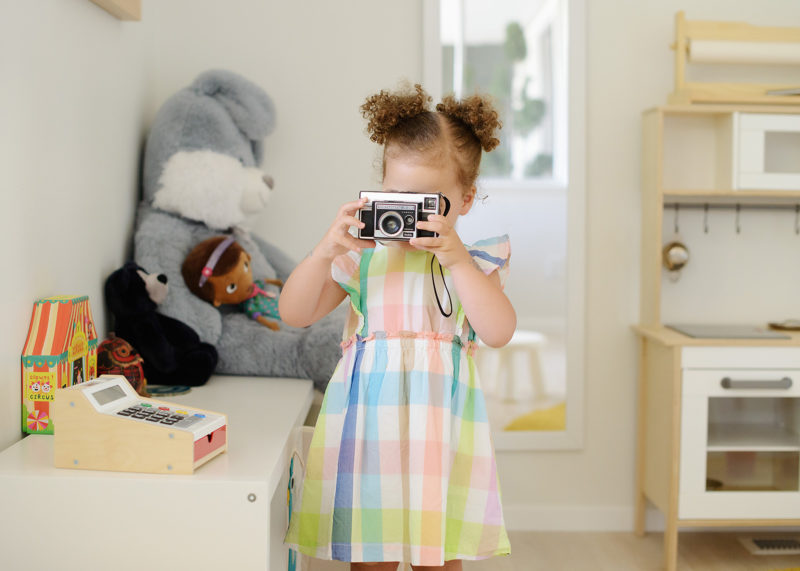 Toddler girl taking a photo in her playroom wearing plaid dress