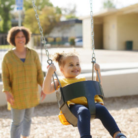 Toddler girl swings in Sacramento outdoor park as mom pushes her