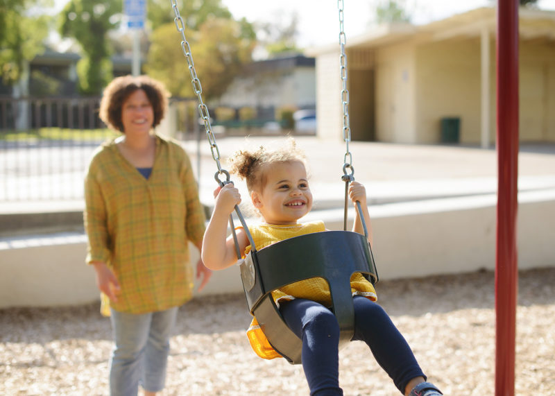 Toddler girl swings in Sacramento outdoor park as mom pushes her