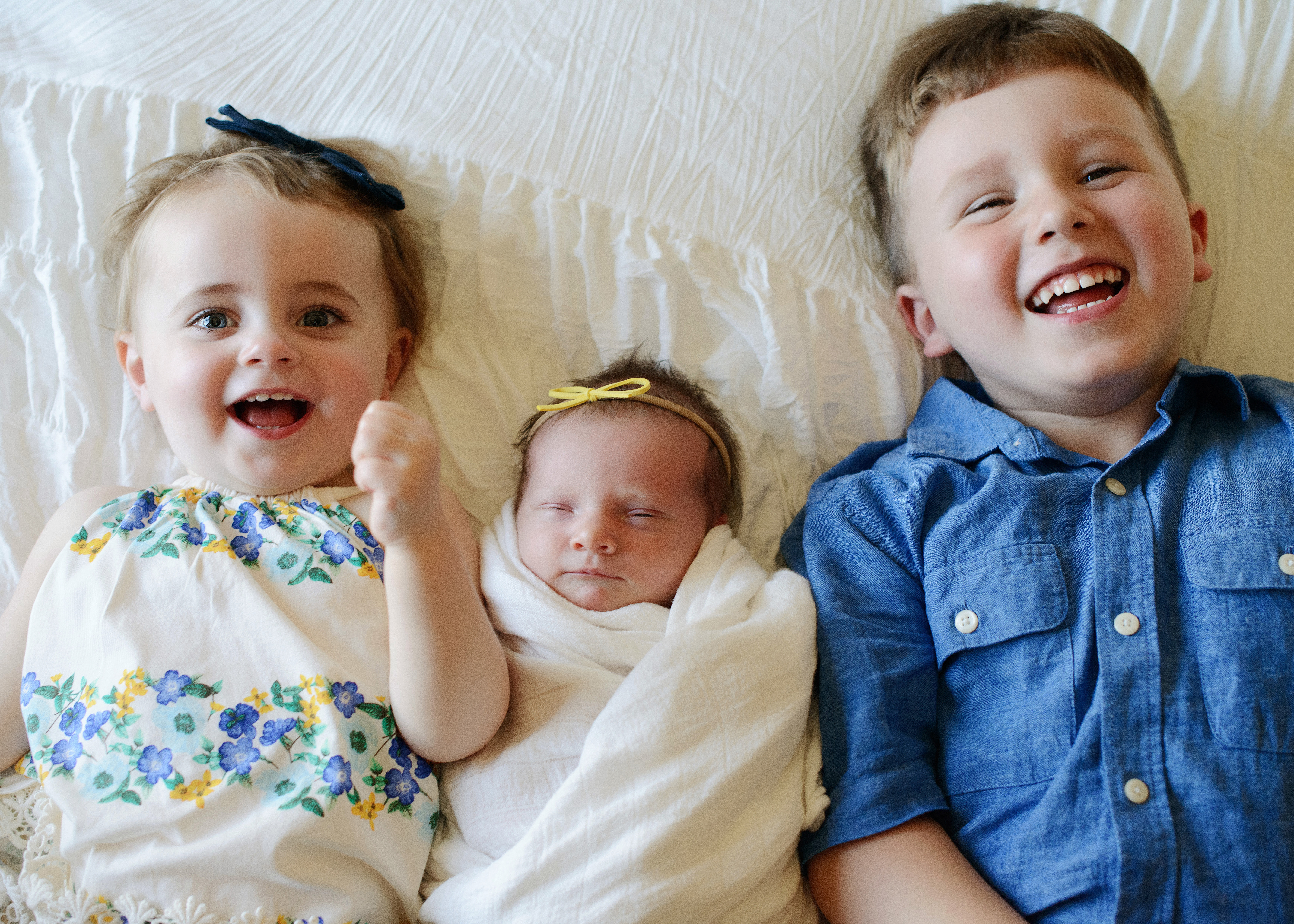 Big sister and brother smile directly into camera as newborn baby sleeps in between them on bed