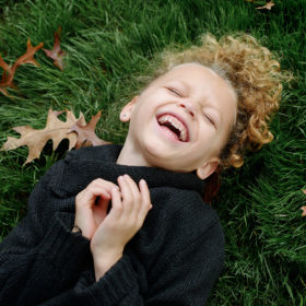 Daughter with blonde curly hair laughing on grass and autumn leaves