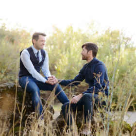 Same sex couple looking lovingly at each other while sitting on a log and holding hands in Fair Oaks