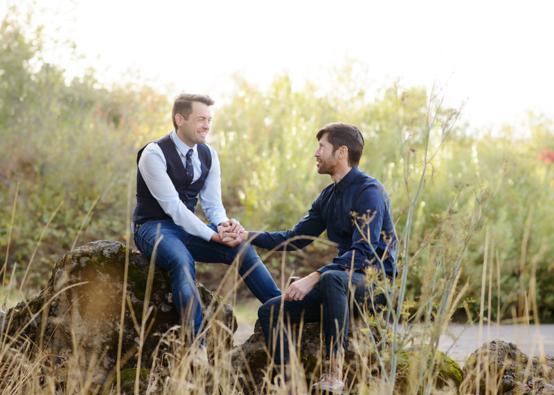 Same sex couple looking lovingly at each other while sitting on a rock and holding hands in Roseville