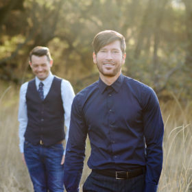 Gay couple posing and smiling for engagement photos during golden hour in dry grass in Fair Oaks