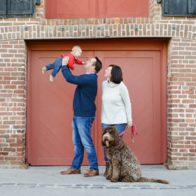 Dad holding baby boy up in the air while mom and dog watch in front of red brick background Old Sacramento
