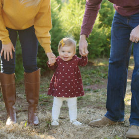 Baby girl standing up while parents hold her hand outdoors