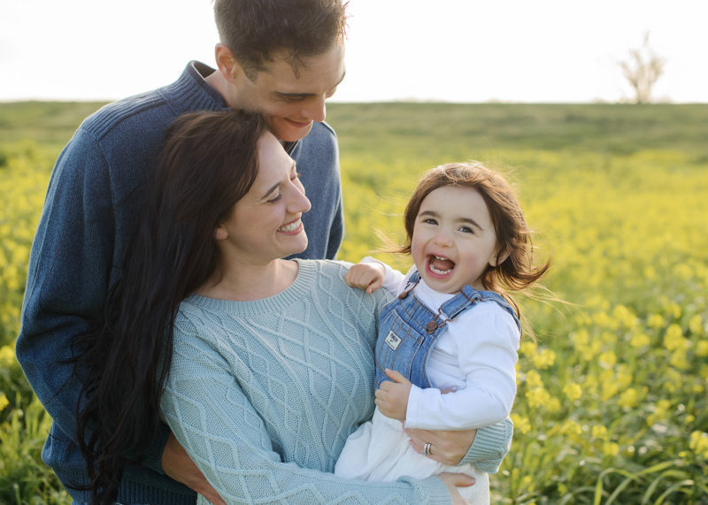 Toddler girl laughing as mom and dad hold her in middle of wildflower field