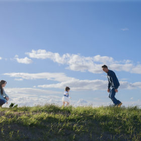 Toddler running towards dad in grassy field with blue sky and clouds