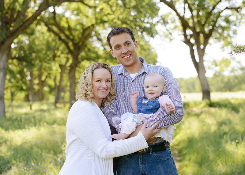 Mom and dad holding baby daughter and smiling with trees and grass background