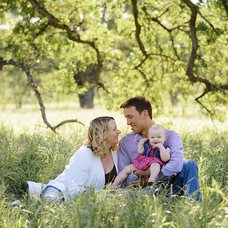 Family portrait with smiling baby girl sitting in grass under a tree in Fair Oaks
