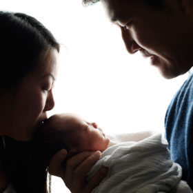 Silhouette of mom and dad kissing newborn baby’s head