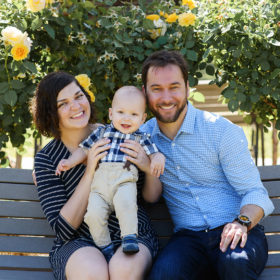 Mom and dad holding baby son on park bench in McKinley Park Rose Garden