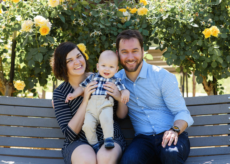 Mom and dad holding baby son on park bench in McKinley Park Rose Garden