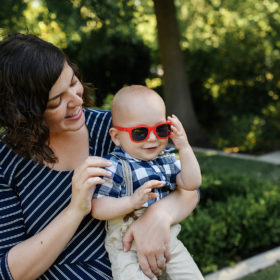 One year old boy wearing red sunglasses whole mom holds him and smiles in Sacramento