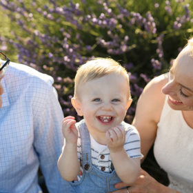 One year old boy laughing and looking directly at camera while mom and dad look lovingly at him in lavender field