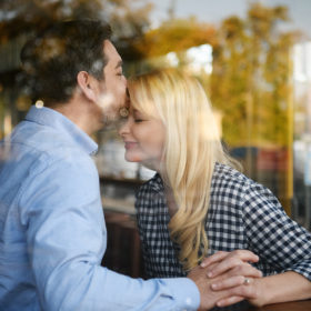Man kissing fiance on forehead in coffee shop with window reflection from outside in Sacramento
