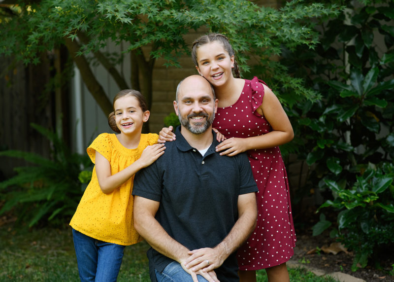 Father and daughters smiling at camera with trees and Fair Oaks home as background
