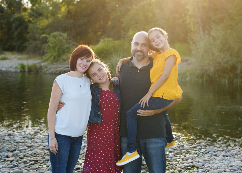 Family photo in natural light with trees and river rocks as background in Fair Oaks