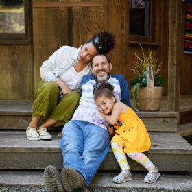 Mom, dad and daughter on backyard steps cuddling with each other in Mendocino home