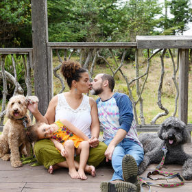 Mom and dad kiss while daughter and dogs look directly at camera while sitting on outdoor deck in Mendocino park