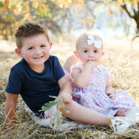 Big brother and baby sister sit on dry grass smiling in Folsom