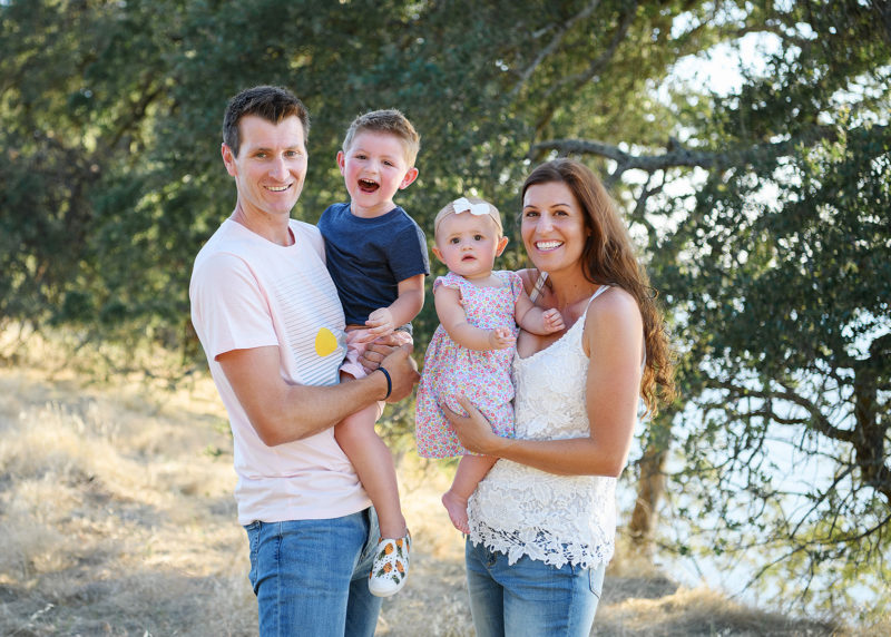 Family portrait with dad holding son and mom holding baby daughter outdoors by trees in Folsom