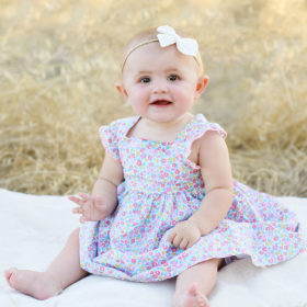Baby girl wearing a bow and floral print dress sitting on blanket outdoors in Folsom