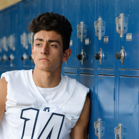 Senior football player in white jersey leaning against blue lockers in Oak View High School El Dorado Hills
