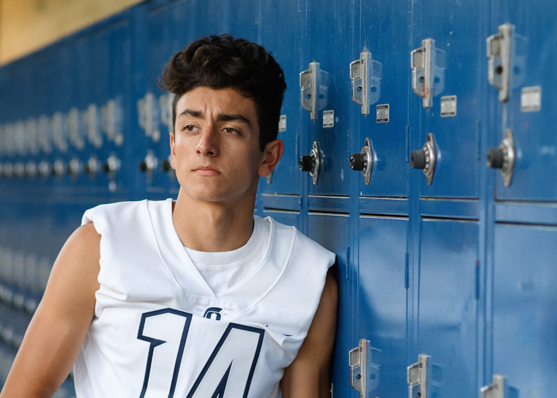 Senior football player in white jersey leaning against blue lockers in Oak Ridge High School El Dorado Hills