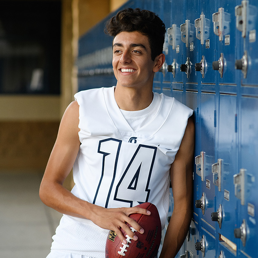 High school senior boy holding a football and wearing a jersey in front of blue lockers at Oak View High El Dorado Hills
