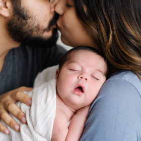 Mom and dad kiss at newborn baby sleeps soundly in their arms in studio