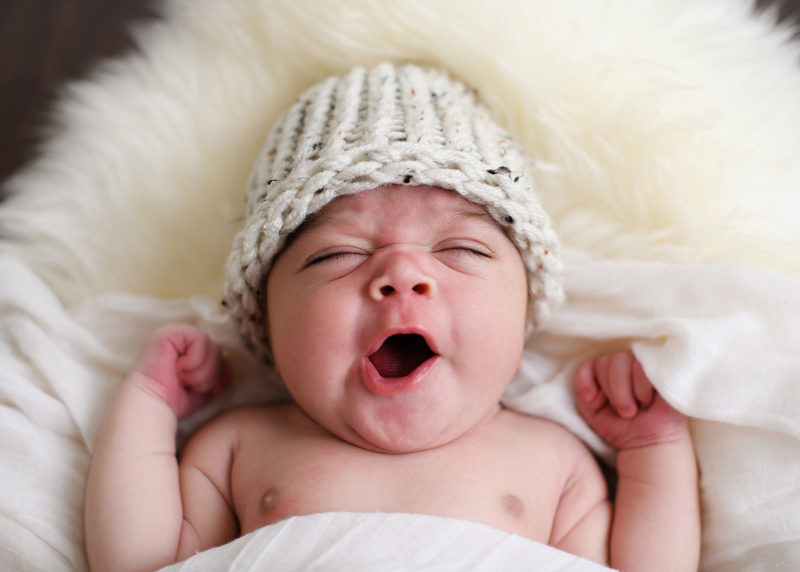 Sleepy newborn baby yawning and wearing a knit beanie on sheepskin rug in studio