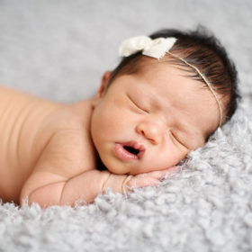 Newborn baby girl sleeping with mouth open with bow on gray blanket in Sacramento studio