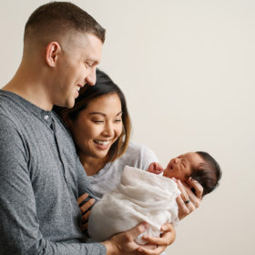 Mom and dad smiling while newborn baby daughter smiles in her sleep in Sacramento studio