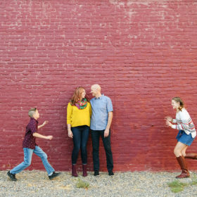 Brother and sister running towards each other as mom and dad look at each other lovingly in front of red brick wall in Sacramento