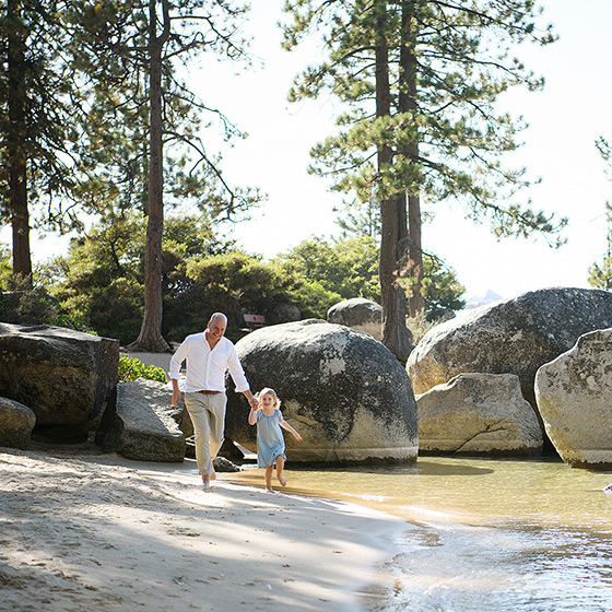 Dad and toddler daughter running on the beach at Lake Tahoe with rocks and trees in background