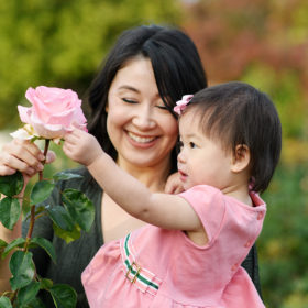 Toddler daughter touching pink rose as mom holds her and smiles at McKinley Park Rose Garden Sacramento