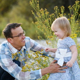 Dad holding toddler daughter by yellow wildflowers and smiling in Fair Oaks