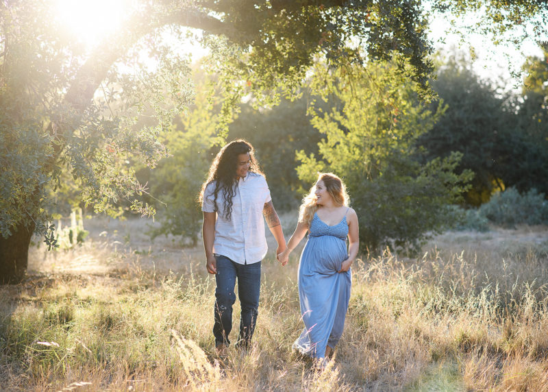 Pregnant woman and dad holding hands and looking at each other under an oak tree