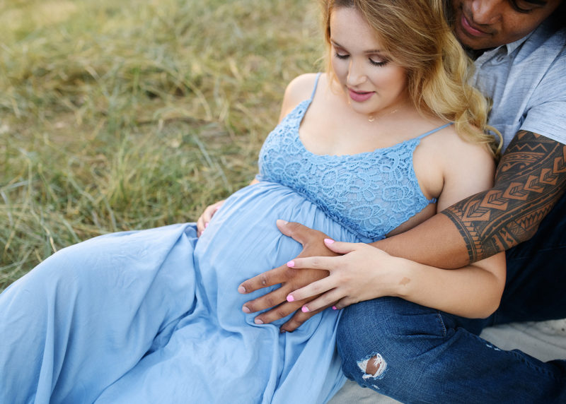 Pregnant woman and man touching baby bump while sitting on grass in Davis