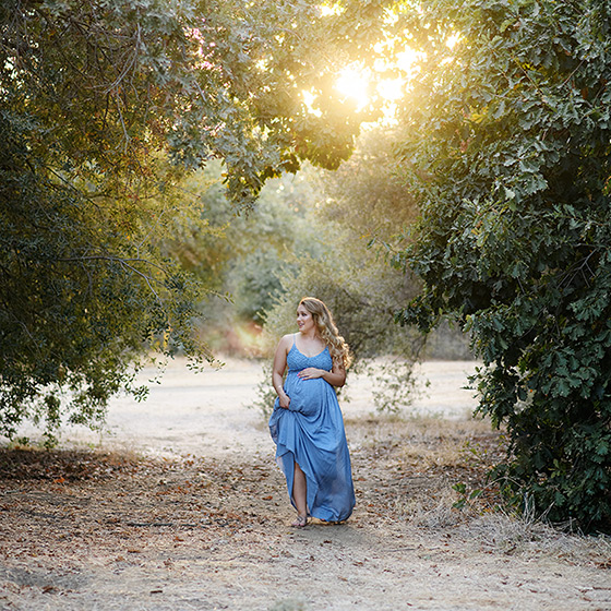 Pregnant woman wearing light blue dress walking in between trees in Davis