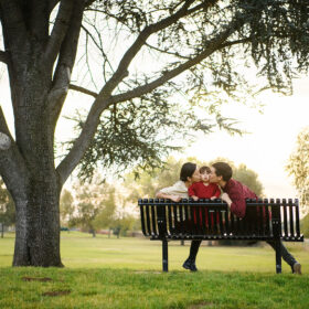 Mom and dad kissing toddler girl on cheek while sitting on a park bench under a tree Rancho Cordova Sacramento