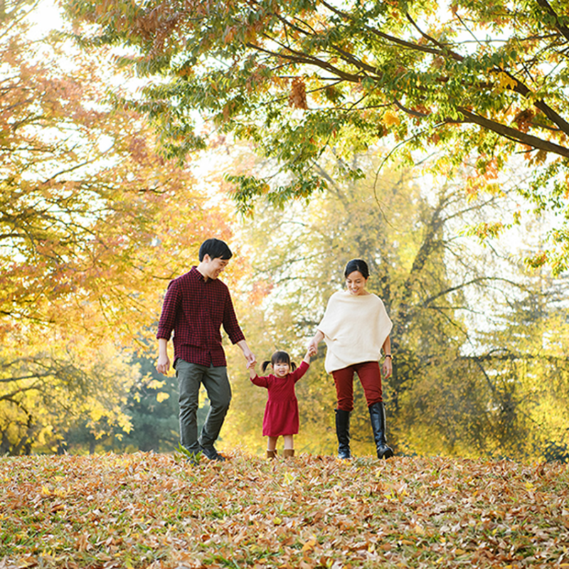 Mom and dad holding toddler girl's hand as they walk through fall leaves